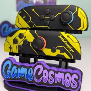 Puede incluir: Mandos Joy-Con de Nintendo Switch negros y amarillos con tem&aacute;tica Cyberpunk 2077. Los mandos tienen un dise&ntilde;o de placa de circuito y el texto "2077" y "Cyberpunk 2077". Se muestran en un soporte "Game Cosmos" morado y azul.