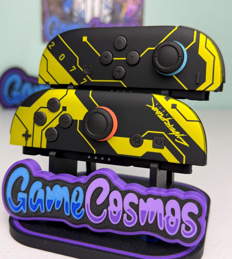 Puede incluir: Mandos Joy-Con de Nintendo Switch negros y amarillos con tem&aacute;tica Cyberpunk 2077. Los mandos tienen un dise&ntilde;o de placa de circuito y el texto "2077" y "Cyberpunk 2077". Se muestran en un soporte "Game Cosmos" morado y azul.
