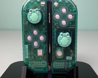 May include: A pair of translucent green Nintendo Switch Joy-Con controllers featuring light pink buttons and custom light green frog-shaped joystick caps. The internal circuitry is visible through the clear green plastic casing.