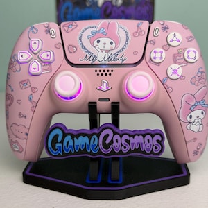 May include: A light pink wireless video game controller featuring My Melody character graphics, cherry icons, and love letters. The controller is mounted on a black stand with a purple and blue GameCosmos logo plate attached to the front base.