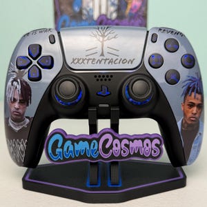 Könnte beinhalten: Ein individueller PlayStation 5-Controller mit einem Porträt von XXXTENTACION, dem Text "XXXTENTACION" und "Game Cosmos" auf dem Ständer. Der Controller hat ein grau-schwarzes Farbschema mit blauen Akzenten.