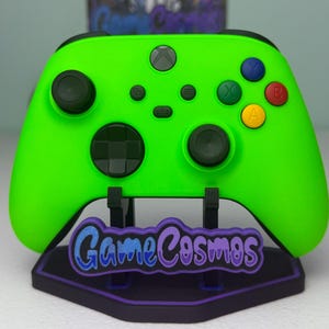 May include: A vibrant green Xbox controller on a black stand, featuring the text "GameCosmos" in blue and purple. The controller has black joysticks and coloured buttons. The stand has a purple border.