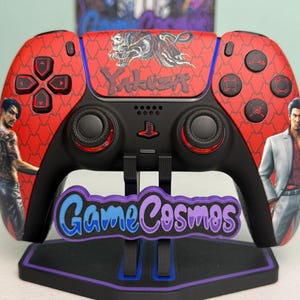 May include: A red and black wireless game controller featuring a dragon graphic and the text Yakuza on the center panel. The controller displays character illustrations on the left and right grips, with black analog sticks and buttons.