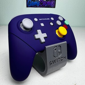 May include: A purple Switchouse controller with a gray stand. The controller has a white plus button, a yellow button, and a green button. The stand has the Nintendo Switch logo and the word "Switch" printed on it.