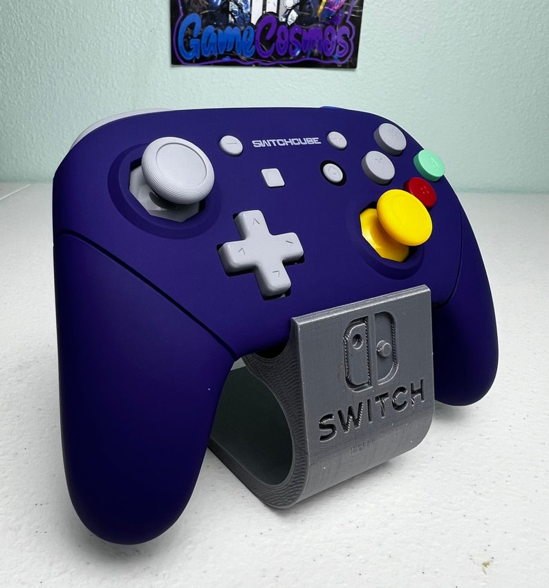 May include: A purple Switchouse controller with a gray stand. The controller has a white plus button, a yellow button, and a green button. The stand has the Nintendo Switch logo and the word "Switch" printed on it.