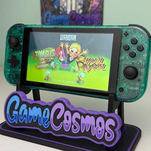 May include: A teal Nintendo Switch console with a screen displaying the "Zombies Ate My Neighbors" game. The console rests on a black stand with the "Game Cosmos" logo in blue and purple.