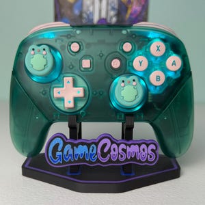 May include: A translucent teal wireless game controller featuring light pink buttons and two analog stick covers shaped like green frogs. The controller is mounted on a black display stand with the text GameCosmos in blue and purple lettering.