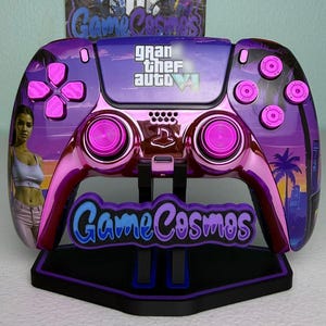 以下が含まれることがあります： 「Game Cosmos」と「Gran Theft Auto VI」の文字が入った、紫とピンクのカスタムビデオゲームコントローラー。 コントローラーの側面には女性の画像があり、黒いスタンドに置かれています。