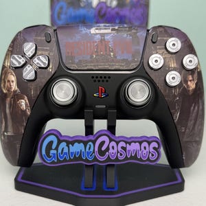May include: A black video game controller featuring Resident Evil graphics, including character portraits and a weapon image. The controller has silver metallic buttons and joysticks, a PlayStation logo, and is displayed on a black stand with a GameCosmos logo.