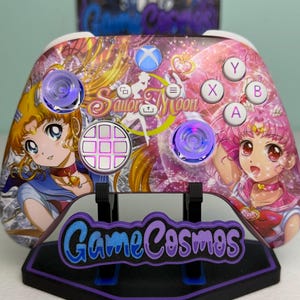 Puede incluir: Un mando de videojuegos inalámbrico con un estampado gráfico de Sailor Moon en tonos rosas y dorados. El mando incluye una cruceta blanca, botones de acción blancos etiquetados como A, B, X, Y y dos joysticks translúcidos sobre un soporte.