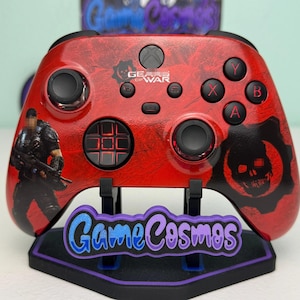 May include: A red wireless video game controller featuring a Gears of War logo and character graphic. The controller has black analog sticks, a directional pad, and action buttons labeled A, B, X, and Y. It rests on a black stand with a GameCosmos logo.