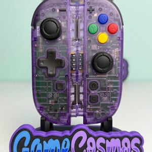 May include: A pair of purple translucent video game controllers with visible internal circuitry, black joysticks, and multicoloured action buttons, mounted on a black stand featuring the text GameCosmos in blue and purple lettering.