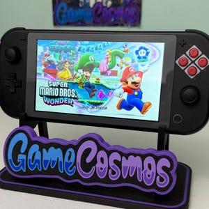 May include: A black handheld gaming console displaying the Super Mario Bros. Wonder game. The console is propped up on a stand with the text "Game Cosmos" in blue and purple. The game features Mario and Luigi.