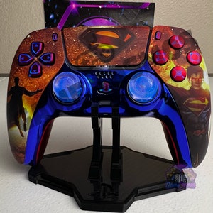 Könnte beinhalten: Ein blauer und roter PlayStation 5-Controller mit Superman-Design. Der Controller steht auf einem schwarzen Ständer. Der Controller hat ein goldenes und rotes Superman-Logo auf der Vorderseite. Der Controller hat blaue und rote Akzente.