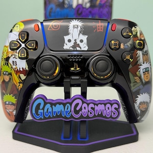 Op de afbeelding: Zwarte PlayStation 5-controller met een aangepast ontwerp met personages uit de Naruto-animeserie. De controller heeft gouden accenten en een standaard met de tekst "Game Cosmos".