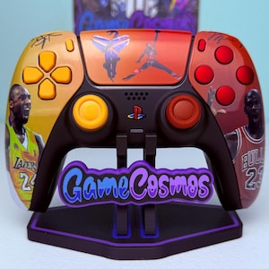 Puede incluir: Mando personalizado de PlayStation 5 con imágenes de las leyendas del baloncesto Kobe Bryant y Michael Jordan. El mando tiene una base negra con detalles en naranja, rojo y amarillo. El mando está sobre un soporte con el texto "GameCosmos".