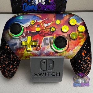 May include: A wireless video game controller featuring a vibrant graphic of a red dragon on the faceplate, yellow action buttons, a yellow directional pad, and black grips with orange paint splatters, resting on a gray plastic stand.