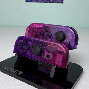May include: Two transparent purple Nintendo Switch Joy-Con controllers are stacked on a black stand. The controllers have black buttons and joysticks. The stand is branded with the Nintendo Switch logo.