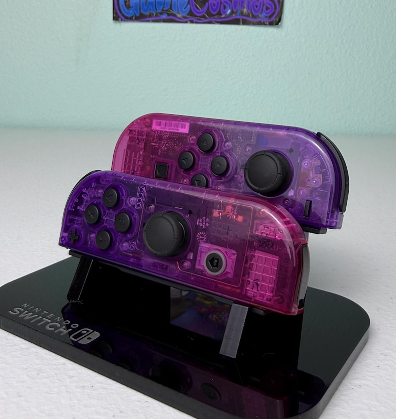 May include: Two transparent purple Nintendo Switch Joy-Con controllers are stacked on a black stand. The controllers have black buttons and joysticks. The stand is branded with the Nintendo Switch logo.