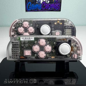 May include: Two Nintendo Switch Joy-Con controllers with transparent shells revealing internal circuitry. Each controller features light pink buttons and a white analog stick. The controllers are mounted on a black Nintendo Switch dock.