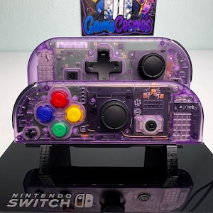 May include: Two purple translucent Nintendo Switch Joy-Con controllers with visible internal circuitry. The front controller features red, blue, yellow, and green action buttons, a black joystick, and a directional pad. The back controller has a black directional pad and joystick.