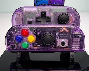 May include: Two purple translucent Nintendo Switch Joy-Con controllers with visible internal circuitry. The front controller features red, blue, yellow, and green action buttons, a black joystick, and a directional pad. The back controller has a black directional pad and joystick.