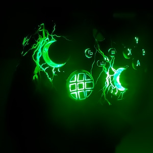 Può includere: Un controller da gioco verde con un design a forma di fulmine e forme di luna crescente e stella che brillano. Il controller ha un pulsante verde con un motivo a griglia.