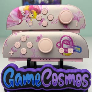 May include: Pink and gray Nintendo Switch Joy-Con controllers with Princess Peach graphics. The controllers have pink buttons and joysticks. The base has the text "Game Cosmos" in blue and purple.