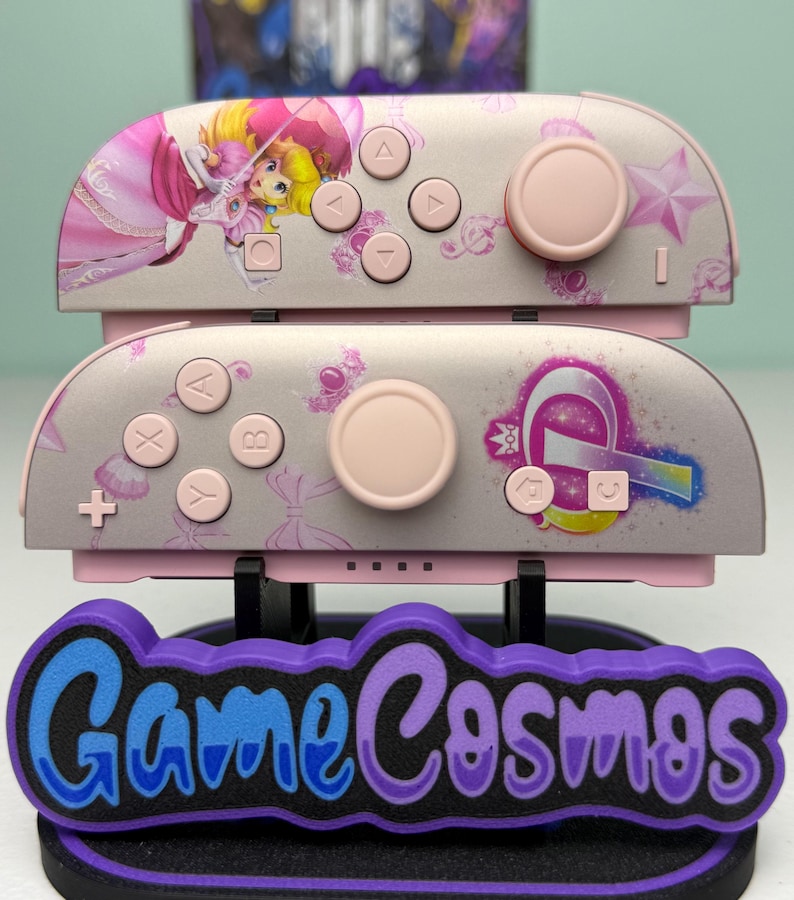 May include: Pink and gray Nintendo Switch Joy-Con controllers with Princess Peach graphics. The controllers have pink buttons and joysticks. The base has the text "Game Cosmos" in blue and purple.