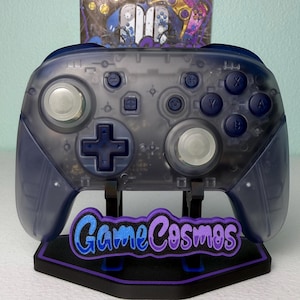 May include: A translucent dark blue wireless video game controller featuring two silver joysticks, a dark blue directional pad, and dark blue action buttons. The controller rests on a black stand with a purple and blue GameCosmos logo.
