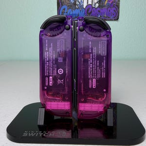 May include: A pair of transparent purple Nintendo Switch Joy-Con controllers, showcasing internal components, are displayed on a black stand. The stand is branded with the Nintendo Switch logo. The controllers are mounted on a black stand.