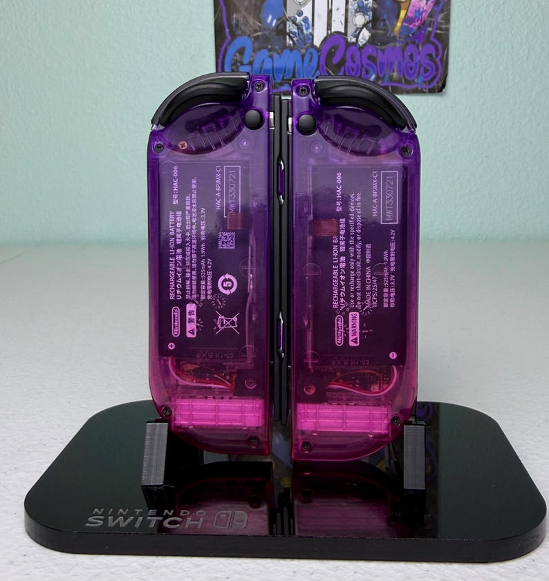 May include: A pair of transparent purple Nintendo Switch Joy-Con controllers, showcasing internal components, are displayed on a black stand. The stand is branded with the Nintendo Switch logo. The controllers are mounted on a black stand.