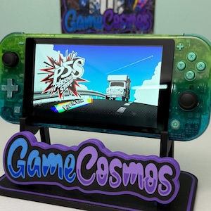May include: A green and blue Nintendo Switch Lite console showing a video game scene. The console is on a black stand with the text "Game Cosmos" in purple and blue. The game's title, "Persona 5 Strikers," is visible on the screen.