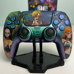 Può includere: Un controller PlayStation 5 personalizzato con personaggi della serie anime One Piece. Il controller presenta una combinazione di colori sfumati teal, viola e blu. Il design include immagini di Sanji, Nami e Robin. Il controller è esposto su un supporto nero.