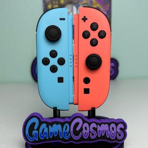 Pu&ograve; includere: Un set di controller Nintendo Switch Joy-Con azzurri e corallo esposto su un supporto. Il supporto presenta il testo "Game Cosmos" in blu e viola. I controller sono in posizione verticale.