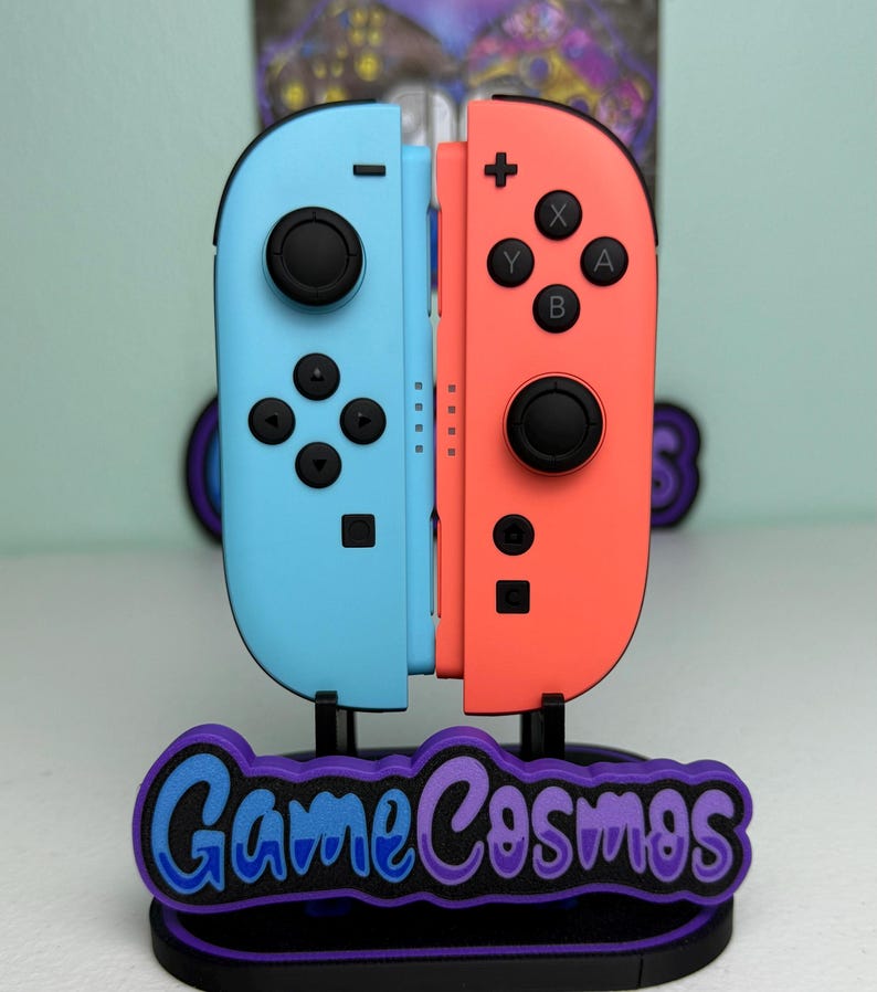 Pu&ograve; includere: Un set di controller Nintendo Switch Joy-Con azzurri e corallo esposto su un supporto. Il supporto presenta il testo "Game Cosmos" in blu e viola. I controller sono in posizione verticale.