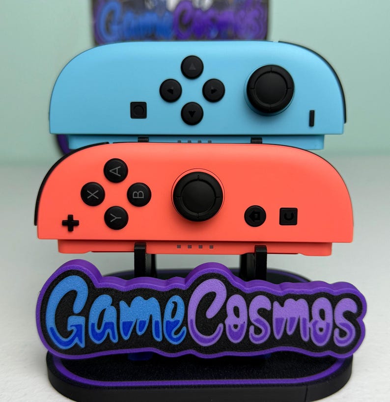 Pu&ograve; includere: Due controller di gioco, uno blu e uno color corallo, impilati su un supporto. Il supporto presenta il testo "Game Cosmos" in blu e viola. I controller hanno pulsanti e joystick neri.