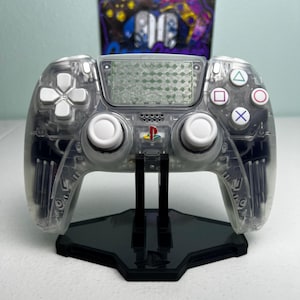 May include: A transparent wireless video game controller with white analog sticks and buttons, featuring a central PlayStation logo and a textured touchpad. The controller is mounted on a black plastic display stand.
