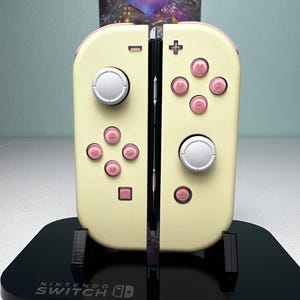 May include: A pair of cream-colored Nintendo Switch Joy-Con controllers featuring light pink buttons and directional pads, with silver analog sticks. The controllers are mounted on a black display stand with the Nintendo Switch logo printed in white.