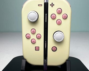 May include: A pair of cream-colored Nintendo Switch Joy-Con controllers featuring light pink buttons and directional pads, with silver analog sticks. The controllers are mounted on a black display stand with the Nintendo Switch logo printed in white.