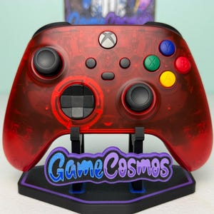 May include: A transparent red Xbox controller with black buttons and a white Xbox logo. The controller is displayed on a black stand with the text "GameCosmos" in blue and purple.