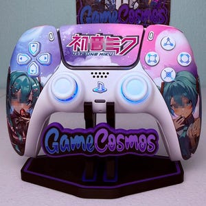 May include: A white wireless video game controller featuring a purple and pink nebula-themed faceplate with Hatsune Miku character graphics and Japanese text. The controller includes blue backlit analog sticks and sits on a black display stand.