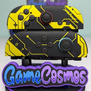 Puede incluir: Dos mandos Joy-Con de Nintendo Switch negros y amarillos con tem&aacute;tica Cyberpunk con un soporte a juego. Los Joy-Cons presentan un dise&ntilde;o de placa de circuito y el texto "Cyberpunk" y "2077". El soporte tiene el texto "Game Cosmos" en azul y morado.