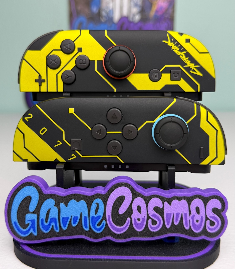 Puede incluir: Dos mandos Joy-Con de Nintendo Switch negros y amarillos con tem&aacute;tica Cyberpunk con un soporte a juego. Los Joy-Cons presentan un dise&ntilde;o de placa de circuito y el texto "Cyberpunk" y "2077". El soporte tiene el texto "Game Cosmos" en azul y morado.