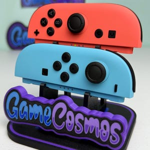 Pu&ograve; includere: Un supporto per controller di videogiochi con un controller rosso e uno blu. Il supporto presenta il testo "Game Cosmos" in blu e viola. I controller hanno pulsanti e joystick neri.