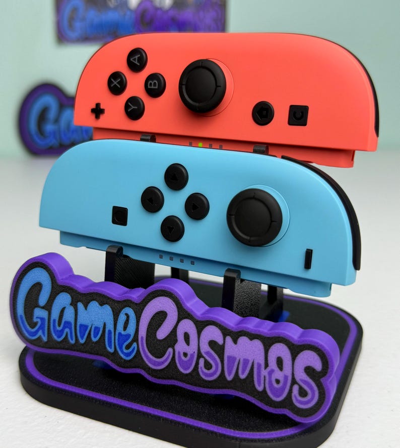Pu&ograve; includere: Un supporto per controller di videogiochi con un controller rosso e uno blu. Il supporto presenta il testo "Game Cosmos" in blu e viola. I controller hanno pulsanti e joystick neri.