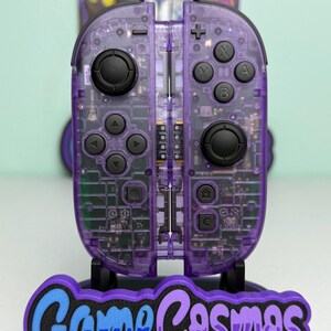 May include: A pair of translucent purple video game controllers with visible internal circuitry, black joysticks, and buttons. The controllers are mounted on a black stand featuring the text GameCosmos in blue and purple lettering.