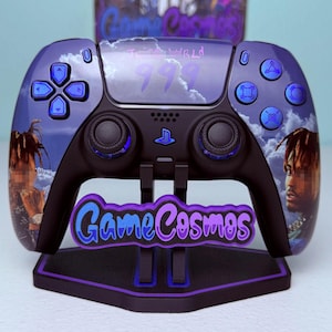 May include: A custom purple and black wireless video game controller featuring a graphic of Juice WRLD, the number 999, and cloudy sky imagery. The controller is mounted on a black stand with the purple text GameCosmos displayed on the front.