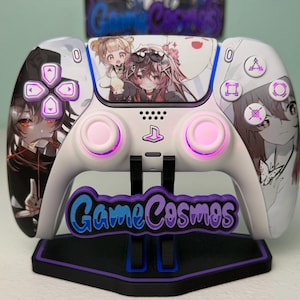 May include: A white wireless video game controller featuring anime-style character illustrations on the front shell. The controller has pink glowing thumbsticks and button icons, resting on a black stand with a purple GameCosmos logo.