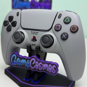 Puede incluir: Un mando gris de PlayStation 5 con botones y joysticks negros, apoyado en un soporte negro y morado. El soporte presenta el texto "Game Cosmos" en azul y morado. El logotipo de PlayStation es visible.